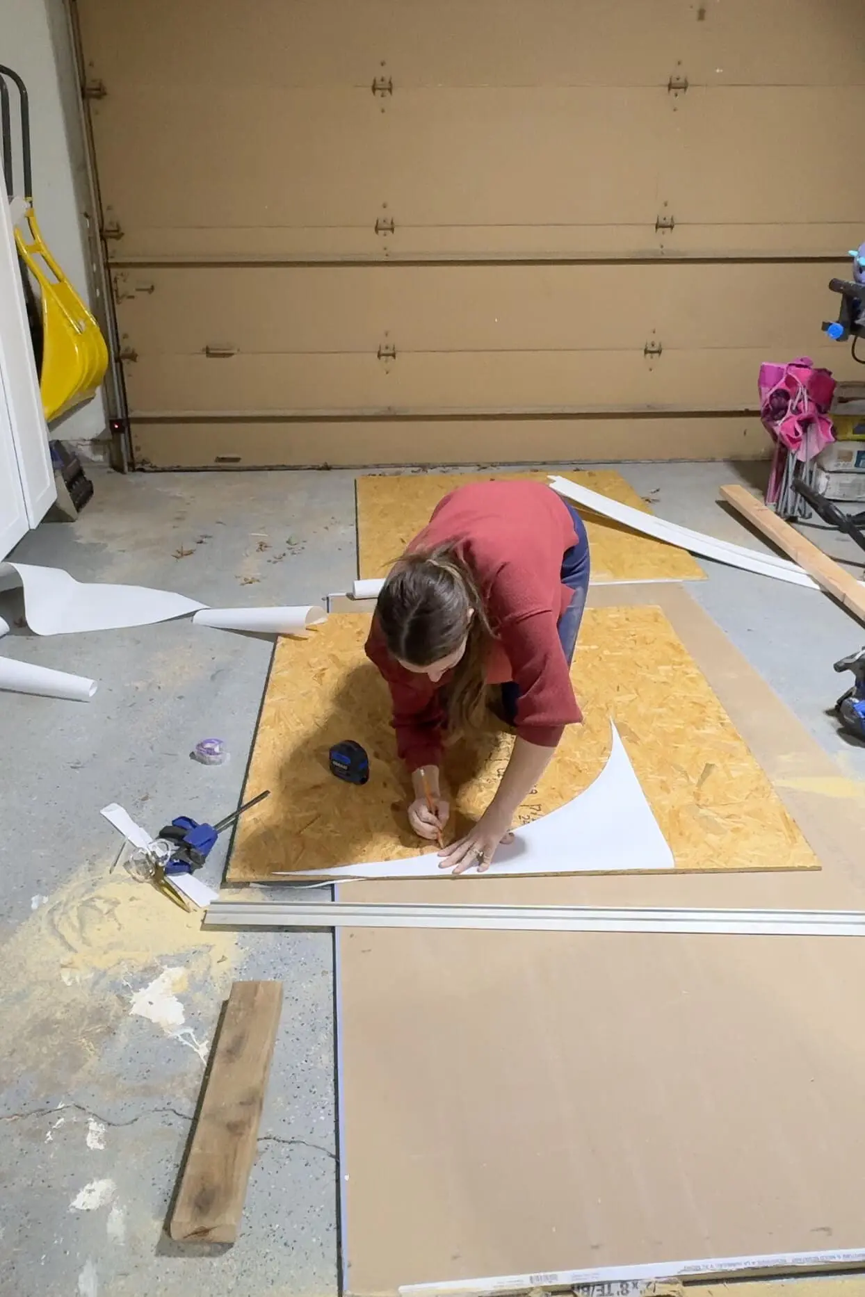 A woman wearing a red sweater and blue jeans leans over a sheet of OSB plywood, carefully tracing an arch shape with a pencil. The garage floor is scattered with tools, wood scraps, and sawdust.